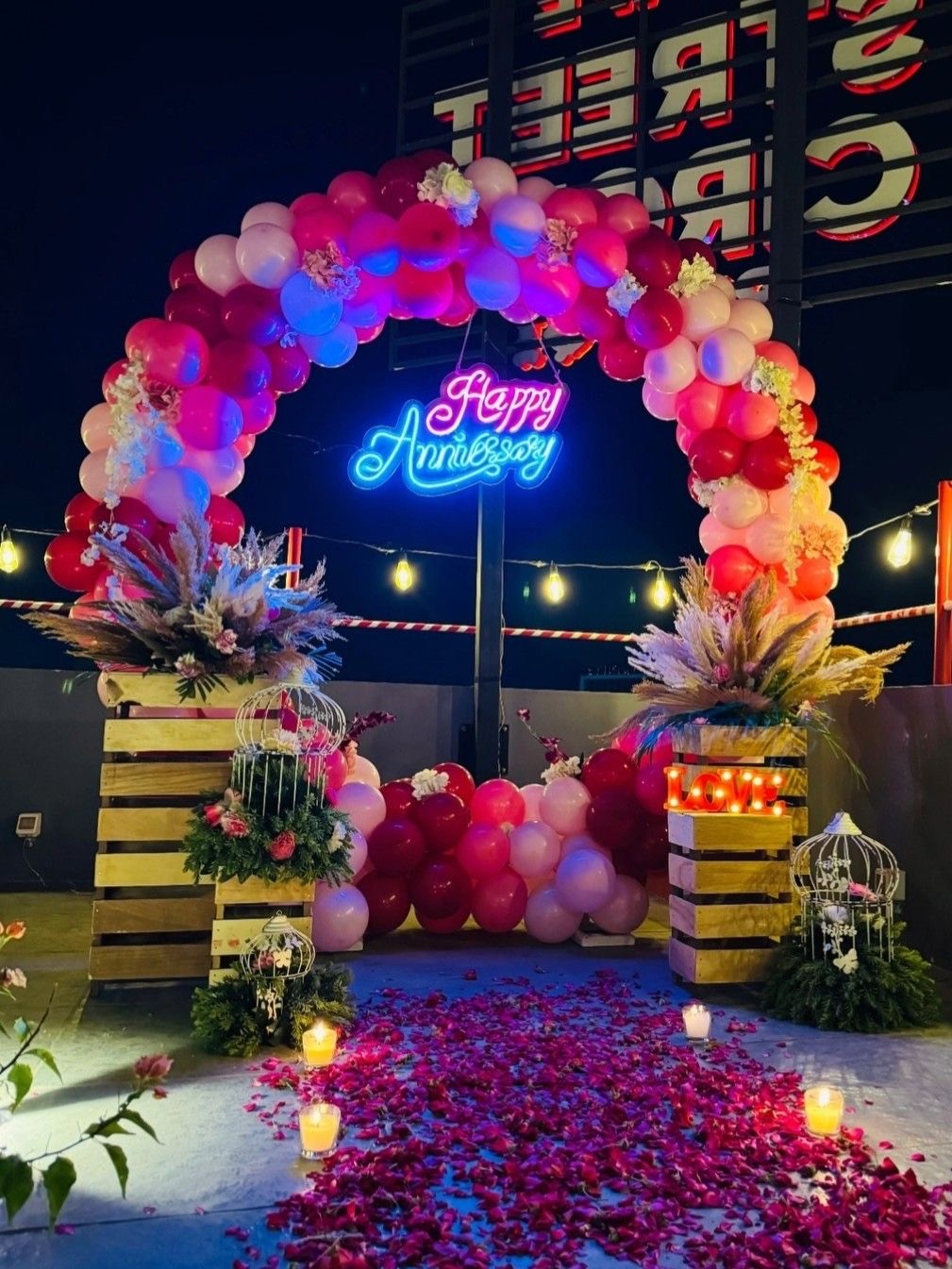 Anniversary decoration in Hyderabad with a pink balloon arch, Happy Anniversary neon sign, candles and rose petals